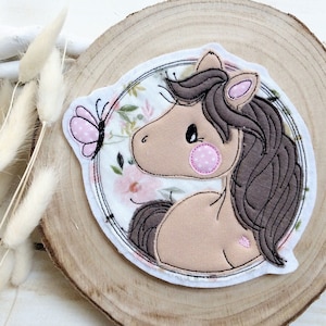 Könnte beinhalten: Ein Stoffaufnäher mit einem Cartoon-Pferdekopf mit brauner Mähne und rosa Akzenten. Das Pferd ist vor einem floralen Hintergrund mit einem rosa Schmetterling platziert. Der Aufnäher befindet sich auf einer Holzscheibe.