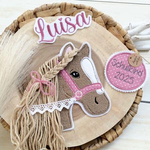 Könnte beinhalten: Handgefertigte Pferde-Dekoration mit einem braunen Pferdekopf mit rosa Halfter und geflochtener Mähne. Die Dekoration enthält den Namen "Luisa" und ein rosa gepunktetes Etikett mit der Aufschrift "Schulkind 2025". Das Pferd ist auf einer Holzscheibe platziert.
