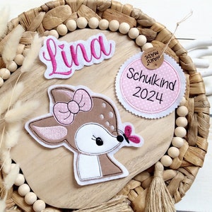 Könnte beinhalten: Eine dekorative Holzplakette mit einem Hirsch-Applikation, einem rosa Namensschild mit "Lina" und einem Etikett mit "Schulkind 2024". Der Hirsch hat eine rosa Schleife und einen Schmetterling. Die Plakette liegt auf einem geflochtenen Tablett.
