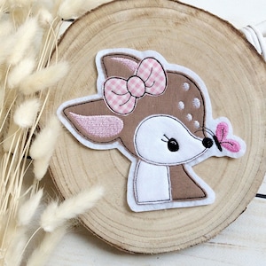 Könnte beinhalten: Ein Hirschaufnäher mit einer rosa Vichy-Schleife und einem rosa Schmetterling. Das Reh hat braunes und weißes Fell mit weißen Flecken. Der Aufnäher befindet sich auf einer Holzscheibe.