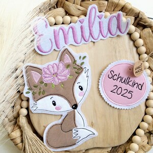 Könnte beinhalten: Gestickte Aufnäher mit einem Fuchs mit Blumenkranz, dem Namen "Emilia" und einem "Schulkind 2025"-Etikett. Der Fuchs ist braun, weiß und rosa, mit grünen Akzenten. Auf einer Holzoberfläche mit geflochtenem Rand.