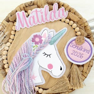 Könnte beinhalten: Runde Holzdekoration mit einem weißen Einhorn, glitzerndem Horn und pastellfarbener Garnmähne. Über dem Einhorn steht der Name "Matilda", und ein Anhänger trägt die Aufschrift "Schulkind 2026". Die Dekoration befindet sich auf einem gewebten Hintergrund.