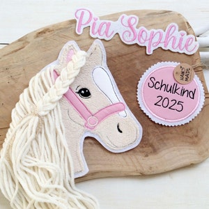 Könnte beinhalten: Beige Pferdekopf-Applikation mit rosa Zaumzeug und langer, cremefarbener Mähne. Die Applikation befindet sich auf einer Holzoberfläche, mit dem Namen "Pia Sophie" in rosa Schrift darüber und einem rosa-weißen "Schulkind 2025" Aufnäher.