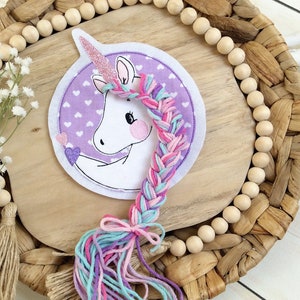 Könnte beinhalten: Ein skurriles Einhorn-Patch mit einer rosa Glitzerhorn und einer geflochtenen Mähne aus rosa, lila und blauem Garn. Das Einhorn ist vor einem lila Hintergrund mit weißen Herzen platziert. Das Patch liegt auf einer Holzoberfläche mit dekorativen Perlen.