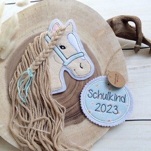 Könnte beinhalten: Applikation eines hellbraunen Pferdekopfes mit geflochtener Mähne und hellblauer Trense. Ein rundes Etikett mit dem Text "Schulkind 2023" ist angebracht. Das Pferd befindet sich auf einem Holzgrund.