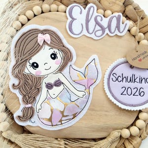 Könnte beinhalten: Ein Filz-Applikation mit Meerjungfrau-Motiv und dem Namen "Elsa" darüber. Die Meerjungfrau hat braunes Haar, eine rosa Schleife und ein lila Muscheloberteil. Ein Anhänger lautet "Handmade with love" und ein runder Aufnäher "Schulkind 2026".