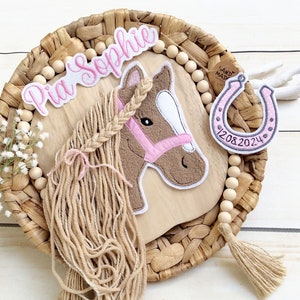 Könnte beinhalten: Ein dekorativer Pferdekopf mit geflochtener Mähne, rosa Zaumzeug und dem Namen "Pia Sophie" in rosa Schrift. Ein Hufeisen mit dem Datum "12.08.2024" ist ebenfalls enthalten. Das Pferd befindet sich auf einer Holzplatte.