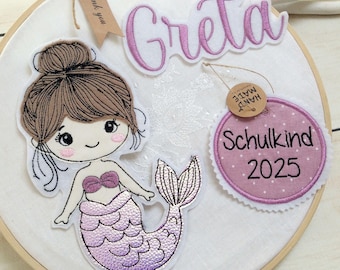 Puede incluir: Un aro de tela blanco con un bordado de sirena y el nombre "Greta" en rosa. Las etiquetas dicen "Thank you" y "Schulkind 2025". La sirena tiene cabello castaño y una cola morada.