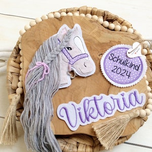 Könnte beinhalten: Dekorative Holzplakette mit einem Plüschpferdekopf, grauer Mähne und lila Halfter. Die Plakette trägt den Namen "Viktoria" und ein Etikett mit der Aufschrift "Schulkind 2024". Das Design ist mit Quasten und Holzperlen verziert.
