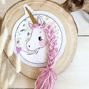 Könnte beinhalten: Ein skurriles Einhorn-Design auf einer Holzscheibe. Das Einhorn hat ein weißes Gesicht, rosa Wangen und ein goldenes Horn. Eine rosa und weiße geflochtene Mähne und ein Schweif aus Garn erstrecken sich vom Kopf des Einhorns. Der Hintergrund hat ein Blumenmuster.
