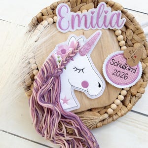 Könnte beinhalten: Ein Einhorn-Design mit einer geflochtenen Mähne aus rosa und lila Garn, einem glitzernden Horn und dem Namen "Emilia" darüber. Ein Etikett lautet "Schulkind 2026". Das Design befindet sich auf einer Holzbasis in einem geflochtenen Korb.