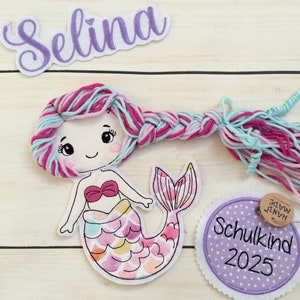 Könnte beinhalten: Ein skurriles Meerjungfrau-Applikation mit einem Zopf aus rosa, blauen und lila Garn. Die Meerjungfrau hat eine rosa Schleife und einen bunten Schwanz. Ein lila Namensschild mit "Selina" und ein Etikett mit "Schulkind 2025" sind ebenfalls vorhanden.