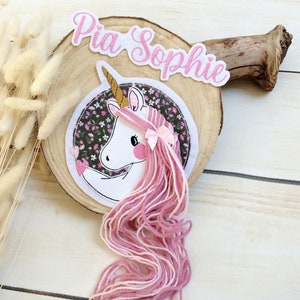 Könnte beinhalten: Ein skurriles Einhorn-Design mit rosa Garnhaar, einem goldenen Horn und einer rosa Schleife. Das Einhorn befindet sich vor einem floralen Hintergrund und einer Holzscheibe. Der Name "Pia Sophie" ist darüber gestickt.