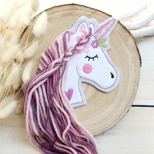 Könnte beinhalten: Ein weißer Einhorn-Aufnäher mit einem rosa Horn und einer fließenden Mähne aus rosa, lila und weißem Garn. Das Einhorn hat einen geflochtenen Haarteil, rosa Wangen und geschlossene Augen. Ein Herz ist auf den Körper des Einhorns gestickt.