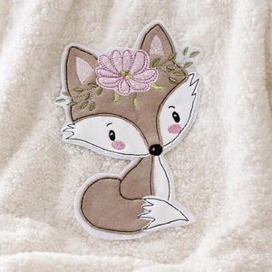 Könnte beinhalten: Eine cremefarbene, texturierte Sherpa-Fleece-Decke mit einer zentralen, gestickten Applikation eines braunen Fuchses, der einen rosa Blumenkranz auf dem Kopf trägt. Der Fuchs hat weiße Akzente an Gesicht und Schwanz sowie rosa Wangen.