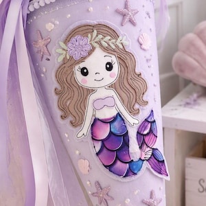 Könnte beinhalten: Eine lavendelfarbene Schultüte mit einer Meerjungfrauen-Applikation mit braunem Haar und violettem Schwanz. Die Oberseite ist mit transparentem violettem Tüll, Bändern und einer weißen Muschel verziert. Der Körper zeigt kleine Seesterne und Perlen.