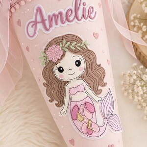 Könnte beinhalten: Eine hellrosa Schultüte mit dem Namen Amelie in Schreibschrift, verziert mit einer passenden rosa Tüllschleife. Auf der Vorderseite befindet sich eine gestickte Meerjungfrau mit braunem Haar und rosa Schwanz, akzentuiert durch kleine rosa Herzen und weiße Perlen.