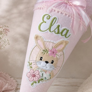 Könnte beinhalten: Eine hellrosa Schultüte mit dem in Grün gestickten Namen Elsa. Die Tüte ist mit einer Applikation eines Hasen mit einer rosa Blume, einem rosa Tüllabschluss mit Satinband und einer kleinen rosa Schleife am unteren Ende verziert.