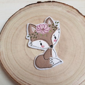 Könnte beinhalten: Gestickter Aufnäher eines Cartoon-Fuchses mit Blumenkranz. Der Fuchs ist braun und weiß mit rosa Wangen und einer schwarzen Nase. Der Blumenkranz ist rosa und grün. Der Aufnäher befindet sich auf einer Holzoberfläche.