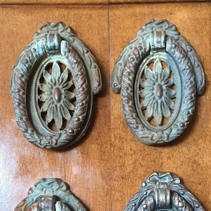 Op de afbeelding: Vier antieke messingkleurige ladegrepen met sierlijke bloemmotieven en een ringvormige handgreep. De grepen hebben een groene patina-afwerking.