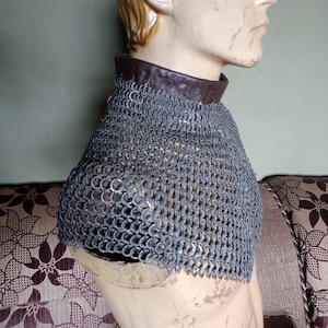 Könnte beinhalten: Ein silberner Kettenhemdkragen mit braunem Lederbesatz. Der Kragen wird von einer Puppe mit einer hellbraunen Perücke getragen.