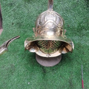 Könnte beinhalten: Ein römischer Gladiatorenhelm aus Messing mit aufwendigen Details. Der Helm hat einen markanten Kamm, einen Gesichtsschutz mit Belüftungslöchern und Wangenpolster. Der Helm wird auf einem grauen Stein vor grünem Hintergrund präsentiert.