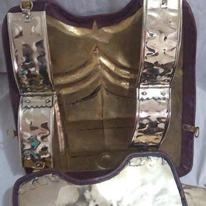 May include: A gold-colored metal breastplate with a decorative design. The breastplate is made of multiple pieces that are joined together. The breastplate is open in the front and back, and the edges are trimmed with a dark brown leather.