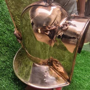 Könnte beinhalten: Eine polierte kupferfarbene Metallskulptur mit reflektierender Oberfläche. Die Skulptur hat eine abgerundete, helmähnliche Form mit einem ausgestellten Sockel und einem gebogenen, flügelartigen Element. Sie steht auf einem roten Sockel vor einem grünen Hintergrund.