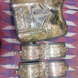 May include: Antique silver-toned metal armour pieces. The set includes a large chest plate and three smaller pieces, possibly for the arms or legs. The metal has a tarnished appearance, suggesting age. The pieces are displayed on a patterned surface.