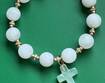 Op de afbeelding: Een kralenarmband met ronde witte stenen kralen afgewisseld met kleine goudkleurige tussenstukjes en heldere kristallen accenten. De armband bevat een lichtgroene stenen kruishanger die in het midden hangt en een goudkleurige karabijnsluiting.