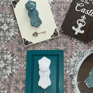 May include: Two decorative door knob hooks with keys, one is teal blue and the other is white. Both are mounted on wooden frames with ornate trim. The teal blue hook is on a white frame and the white hook is on a teal blue frame.
