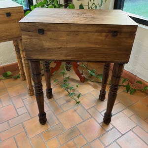 May include: A wooden cabinet on four dark brown, turned legs. The cabinet has a hinged lid and metal clasps. The top of the cabinet has the text "CABALLERO" etched into the wood. The cabinet is set on a terracotta tile floor.
