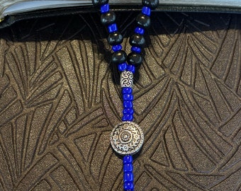 Può includere: Un rosario con perline di legno nere e perline distanziatrici blu. Il rosario presenta un ciondolo a forma di croce color argento e un elemento centrale decorativo color argento. Il rosario è esposto su un libro aperto.
