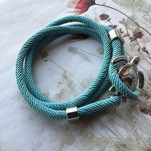 May include: A turquoise blue rope bracelet with a silver turtle charm. The bracelet is made of two strands of rope that are twisted together.