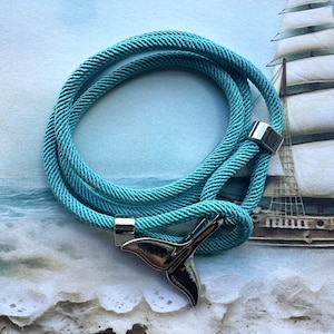 Könnte beinhalten: Ein türkisfarbenes Seil-Armband mit einem silbernen Wal-Schwanz-Anhänger. Das Armband besteht aus einem geflochtenen Seil und hat einen silbernen Verschluss.
