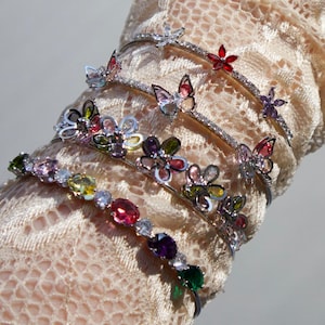 May include: A collection of silver bracelets featuring butterfly and floral designs in various colours. The bracelets are decorated with gemstones in shades of red, pink, green, and yellow. The bracelets are worn on a lace sleeve.