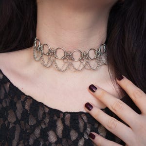 May include: A silver choker necklace with a layered design. The necklace features interconnected rings and dangling chains. The jewellery is worn around the neck and is made of a shiny, metallic material.