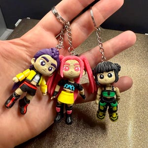 May include: Three keyrings with miniature figures in vibrant colours. Each figure has a distinct hairstyle and outfit, with detailed facial features. The keyrings are attached to silver rings. The figures are in yellow, pink, and green.