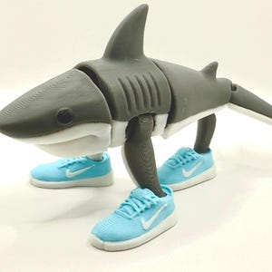 May include: A gray and white articulated shark figure wearing light blue sneakers with white soles and a white swoosh logo. The shark has a dorsal fin and a segmented body. The toy is posed as if walking.