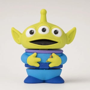 May include: A small three-eyed alien figurine with lime green skin, a pointed antenna on its head, and large pointed ears. The character wears a blue space suit with a purple collar, a dark blue belt, and dark blue boots, with hands resting on its chest.
