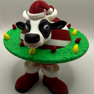 Könnte beinhalten: Eine skurrile Weihnachts-Kuh-Figur. Die Kuh hat ein schwarz-weißes Gesicht, trägt einen Weihnachtsmannhut und ist von einem grünen Kranz mit roten und gelben Akzenten umgeben. Sie trägt rote Stiefel.