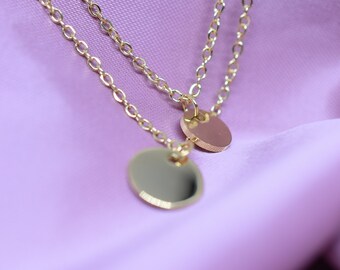 Puede incluir: Un collar de cadena de oro con dos colgantes redondos. El collar está sobre un fondo de tela morado.