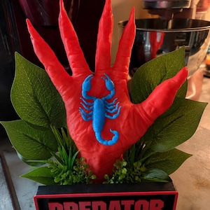 May include: A red hand-shaped sculpture with long, pointed fingers featuring a bright blue scorpion centered on the palm. The sculpture is set in a black rectangular base with the word PREDATOR in red letters, surrounded by green artificial leaves.