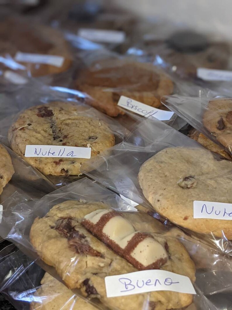 May include: A variety of cookies in clear plastic bags, labeled with their flavors: Nutella, Bueno, and Biscoff. The cookies are arranged in a row, with the labels facing the viewer.