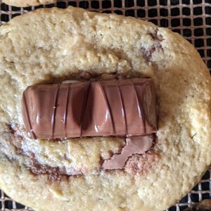 May include: A close-up of a golden brown cookie with a chocolate bar on top. The cookie is on a brown wire rack.