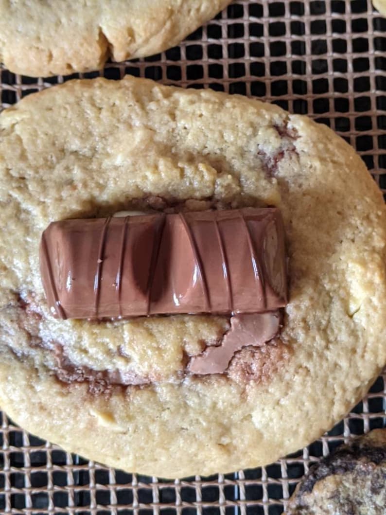 May include: A close-up of a golden brown cookie with a chocolate bar on top. The cookie is on a brown wire rack.