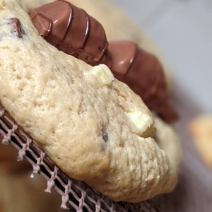 May include: Close-up of a chocolate chip cookie with a chocolate bar on top. The cookie is light brown and has white chocolate chips. The chocolate bar is dark brown and has a white stripe.