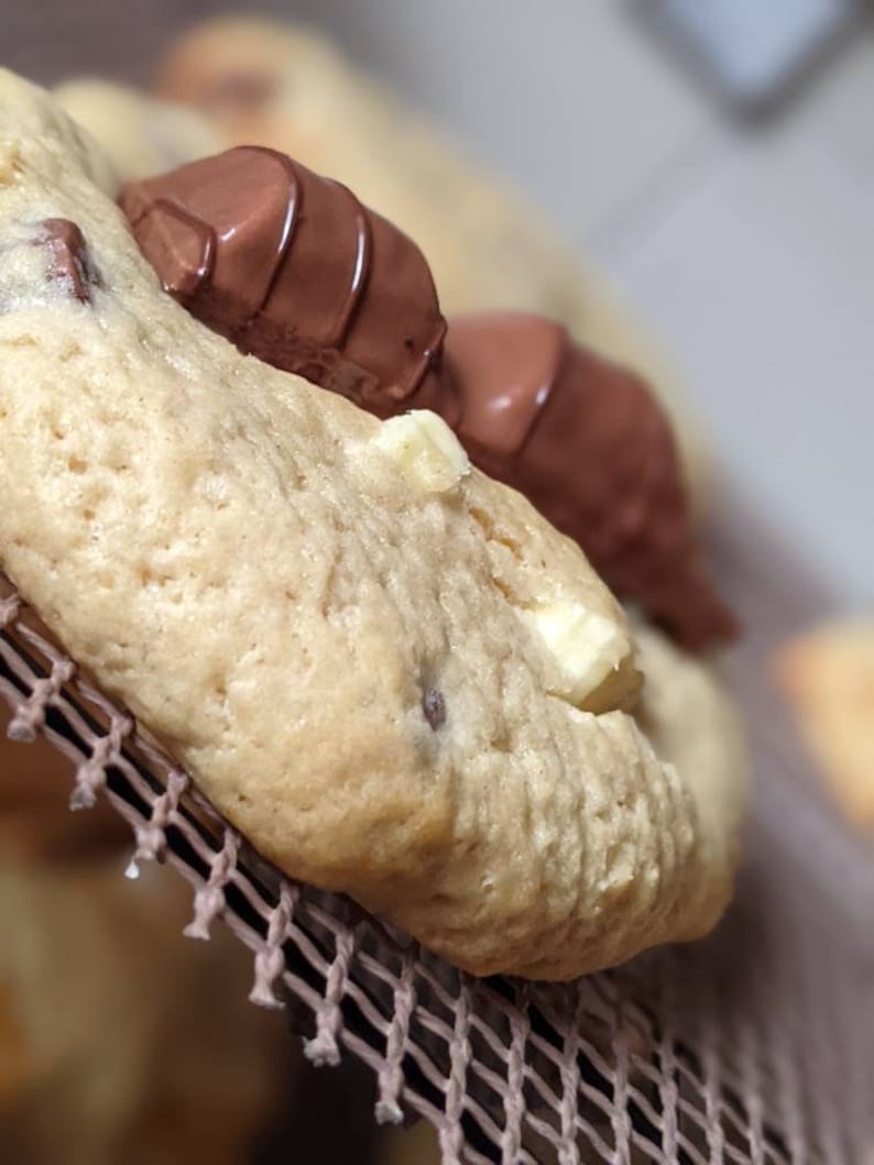 May include: Close-up of a chocolate chip cookie with a chocolate bar on top. The cookie is light brown and has white chocolate chips. The chocolate bar is dark brown and has a white stripe.