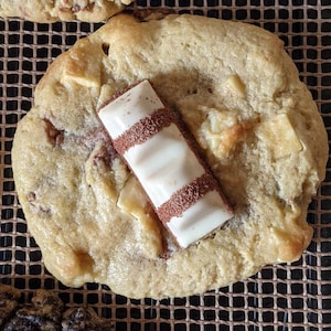 May include: A close-up of a golden brown cookie with white chocolate chips and a white chocolate bar on top. The cookie is on a brown wire rack.