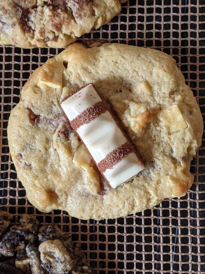 May include: A close-up of a golden brown cookie with white chocolate chips and a white chocolate bar on top. The cookie is on a brown wire rack.